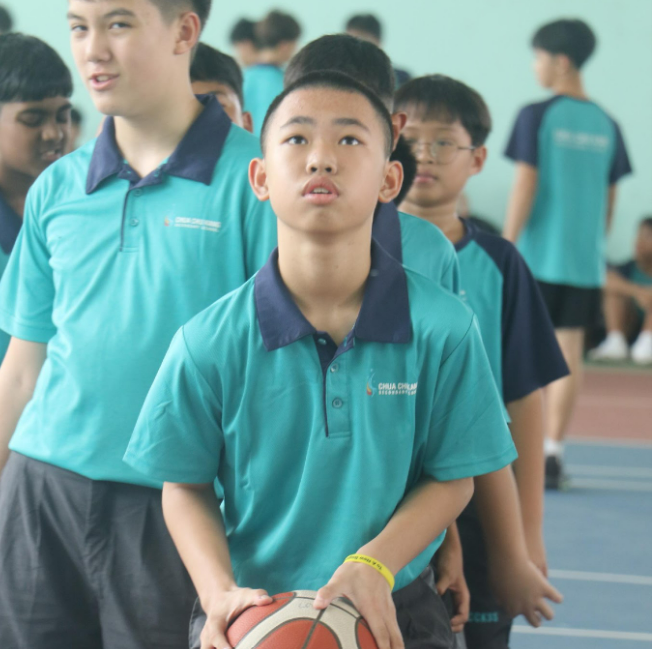 A boy in a teal shirt holds a basketball while several teammates stand behind him on an indoor court.